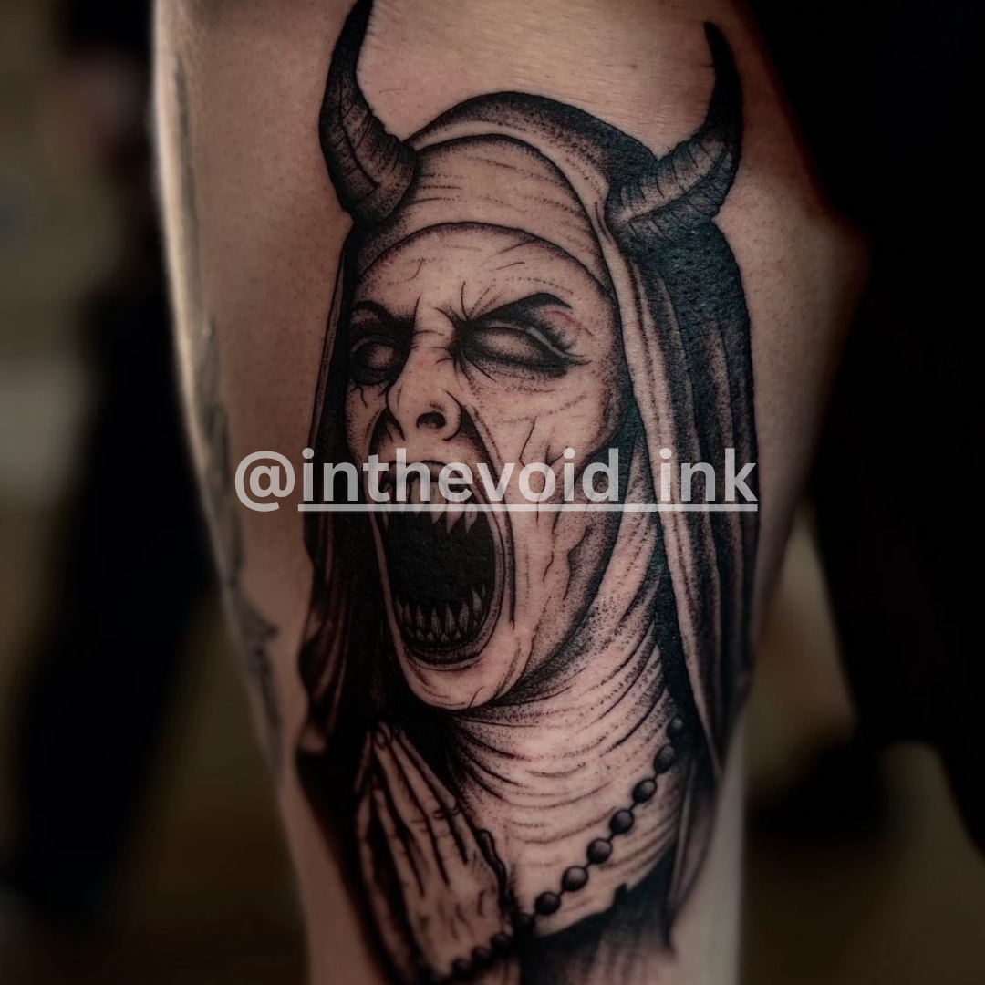 Tattoo of a demonic face with horns on a person's arm, featuring the artist's name '@inthevoid_ink'.