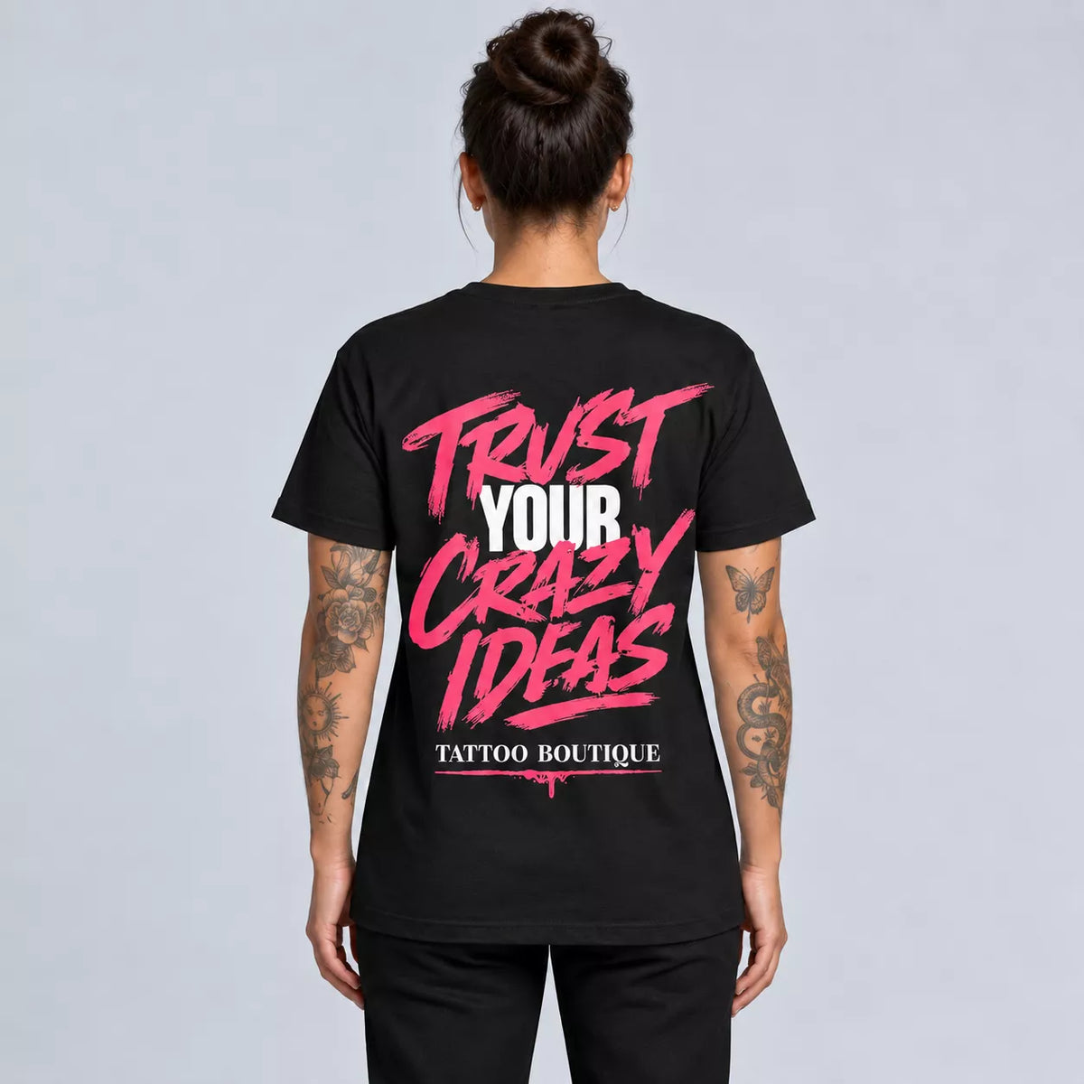 A woman viewed from the back wearing a black short-sleeve T-shirt with a large graphic print on the back. The print reads 'TRUST YOUR CRAZY IDEAS' in pink and white, with 'TATTOO BOUTIQUE' below in smaller white text.