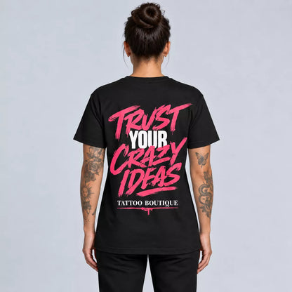 A woman viewed from the back wearing a black short-sleeve T-shirt with a large graphic print on the back. The print reads 'TRUST YOUR CRAZY IDEAS' in pink and white, with 'TATTOO BOUTIQUE' below in smaller white text.