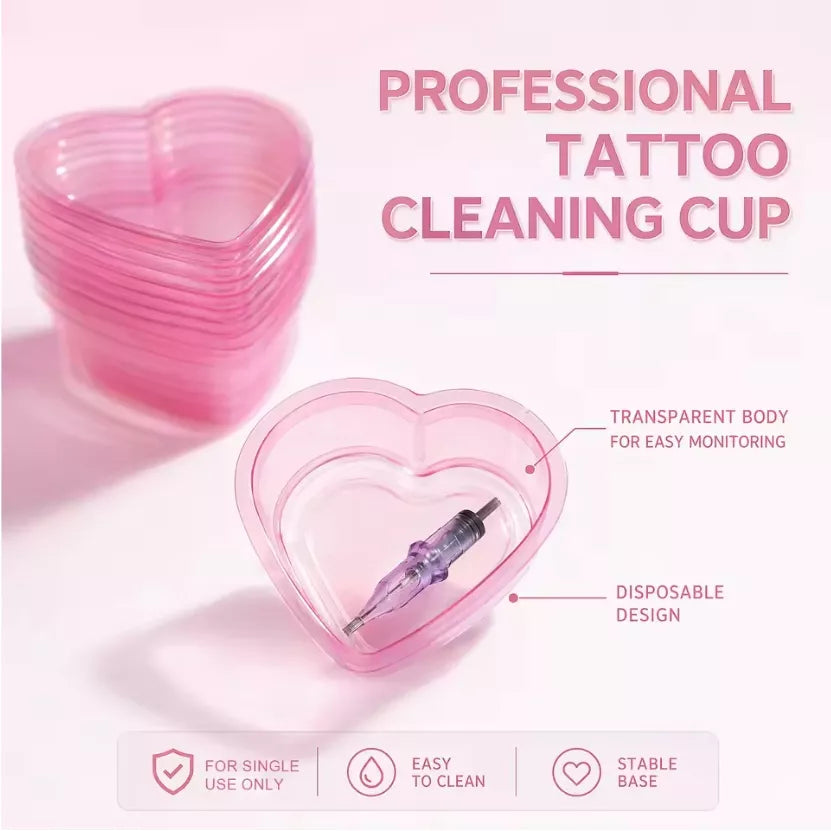 Pink heart-shaped transparent tattoo cleaning cup shown on a light pink background with stacked cups, a syringe inside one cup, and text reading 'PROFESSIONAL TATTOO CLEANING CUP' plus feature callouts.