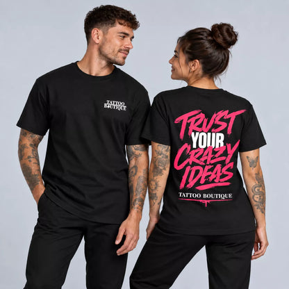 A man and a woman modeling black graphic t-shirts against a light gray studio background. The man wears a black t-shirt with a small white 'TATTOO BOUTIQUE' logo on the left chest.