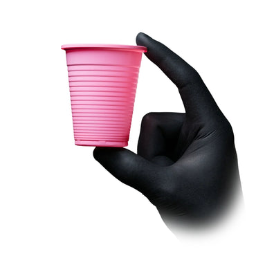A hand wearing a black nitrile glove holds a pink disposable plastic tattoo rinse cup with horizontal ridges against a plain white background.