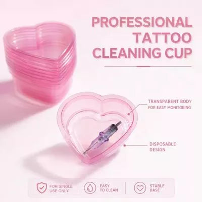Pink heart-shaped transparent tattoo cleaning cup shown on a light pink background with stacked cups, a syringe inside one cup, and text reading 'PROFESSIONAL TATTOO CLEANING CUP' plus feature callouts.
