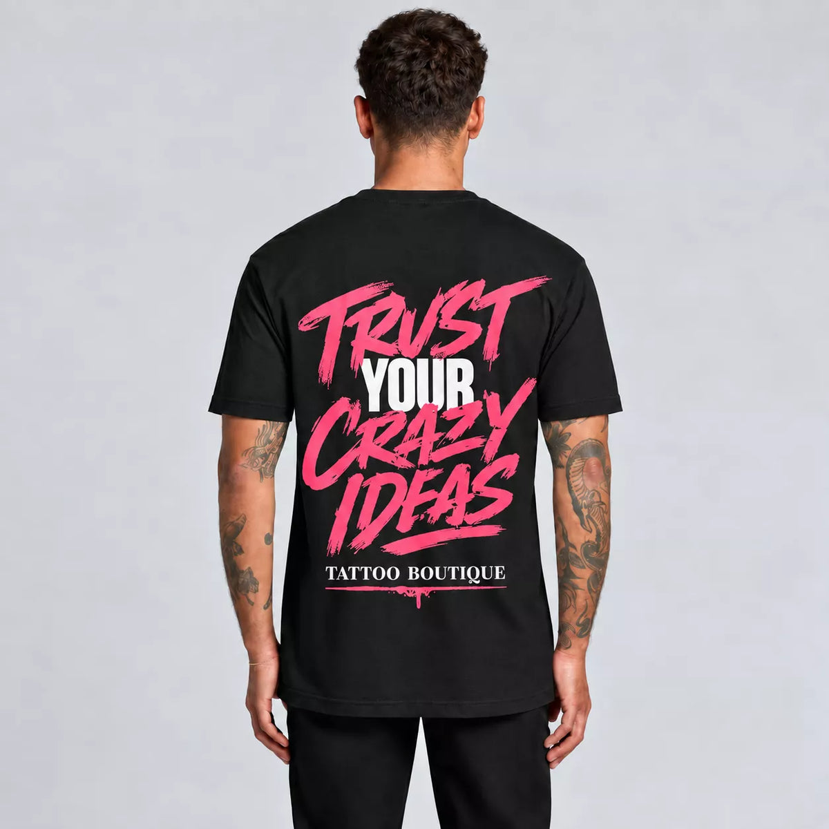A man viewed from behind wearing a black t-shirt with a large graphic on the back reading 'TRUST YOUR CRAZY IDEAS' in pink and white, and 'TATTOO BOUTIQUE' below it, against a light gray studio background.