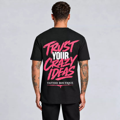 A man viewed from behind wearing a black t-shirt with a large graphic on the back reading 'TRUST YOUR CRAZY IDEAS' in pink and white, and 'TATTOO BOUTIQUE' below it, against a light gray studio background.