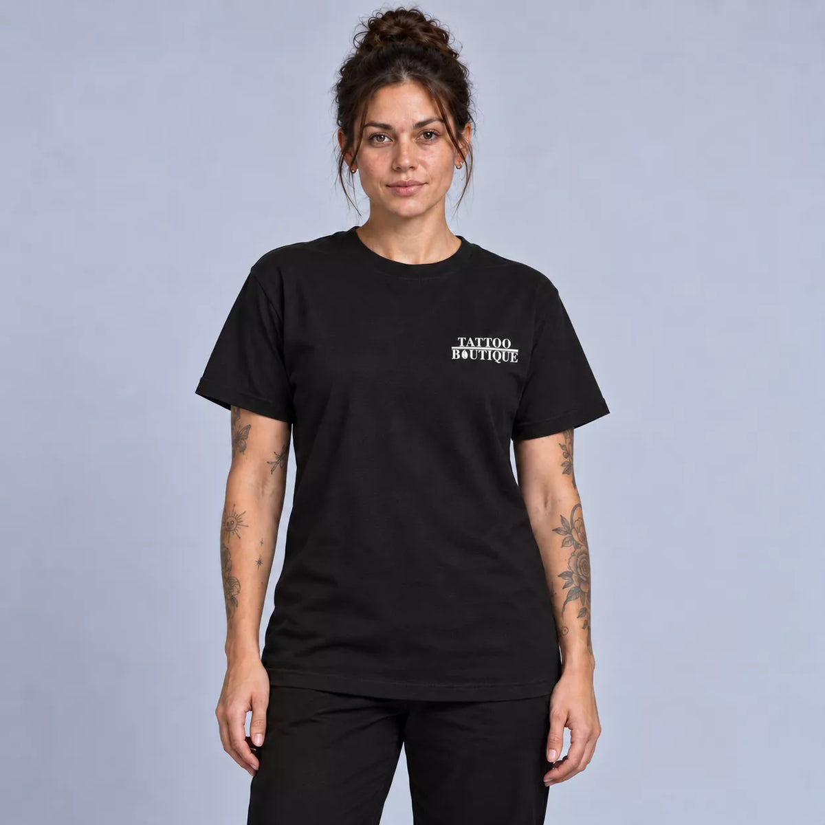A woman modeling a black short-sleeve crewneck t-shirt with 'TATTOO BOUTIQUE' printed in white on the left chest, set against a solid light gray studio background.