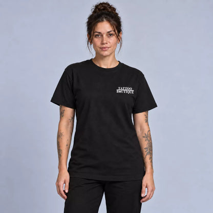 A woman modeling a black short-sleeve crewneck t-shirt with 'TATTOO BOUTIQUE' printed in white on the left chest, set against a solid light gray studio background.