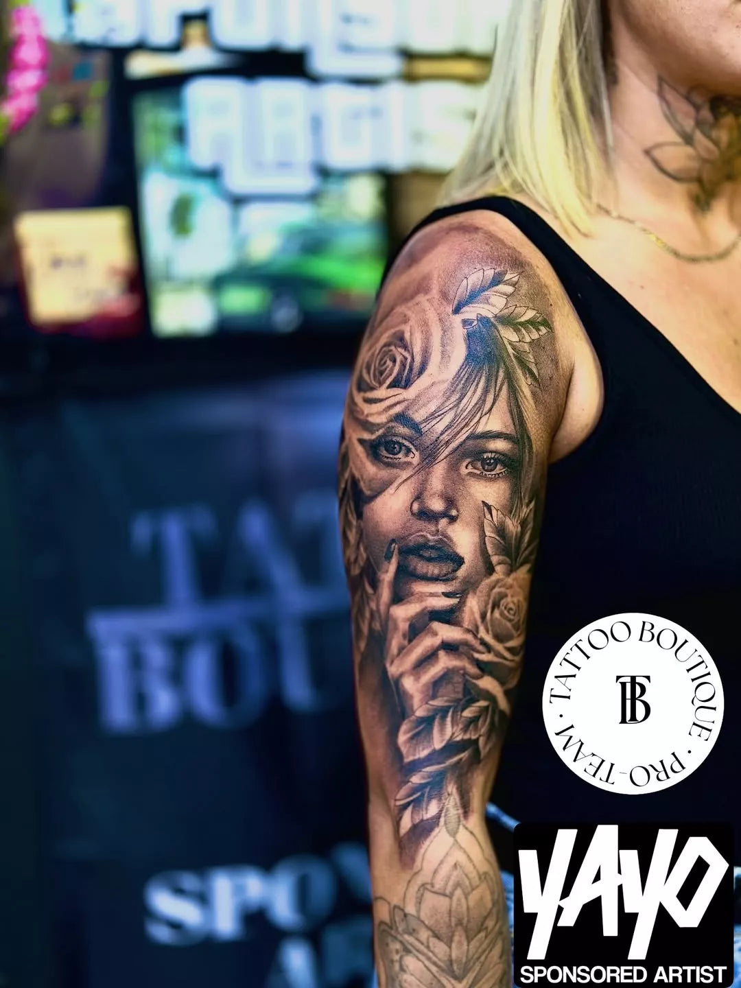 Person with a detailed tattoo of a face on their arm, with 'Tattoo Boutique Pro Team' and 'Yayo' logos in the background.