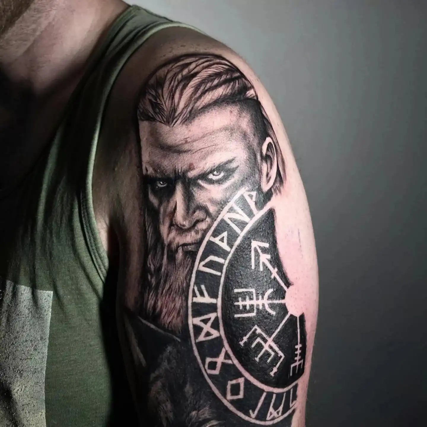 Tattoo of a warrior's face with a shield featuring runes on a person's arm.