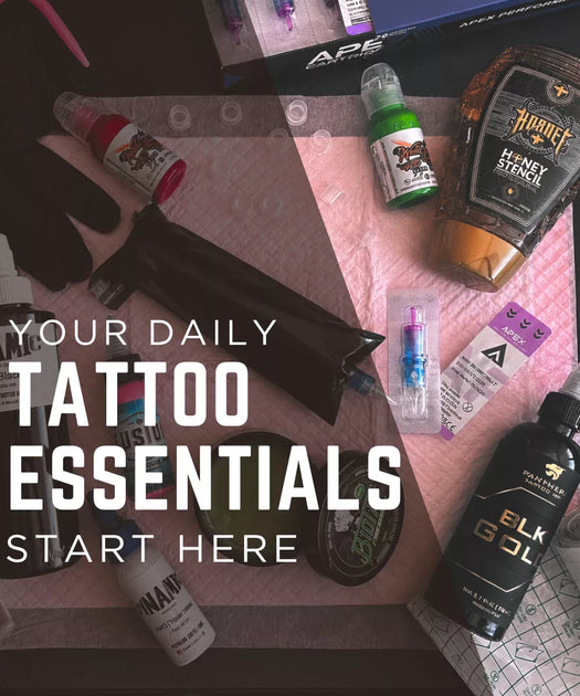 A styled flat-lay of tattoo supplies on a pink patterned mat, including a black tube, gloves, a honey stencil bottle, a small green spray bottle, a black Elk Gold ink bottle, and other tattoo products, with large overlaid text reading 'YOUR DAILY TATTOO ESSENTIALS START HERE'.